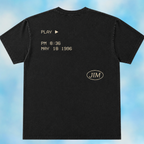 A black short-sleeve t-shirt shown from the back, featuring a light beige retro video game interface design with the text 'PLAY PM 8:36 MAY 18 1996' and a circular 'JIM' logo, set against a blue cloudy background.