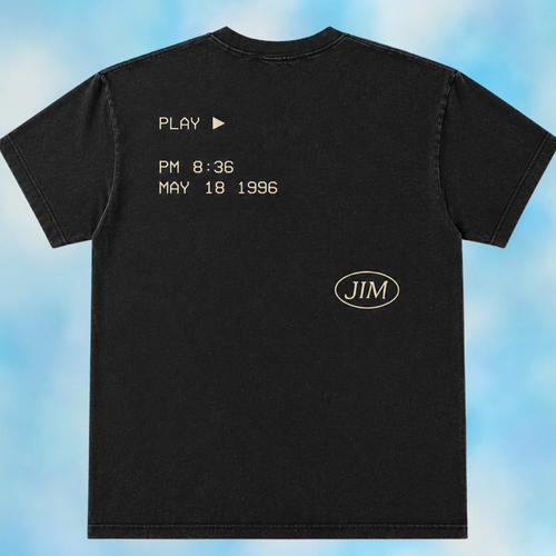 A black short-sleeve t-shirt shown from the back, featuring a light beige retro video game interface design with the text 'PLAY PM 8:36 MAY 18 1996' and a circular 'JIM' logo, set against a blue cloudy background.